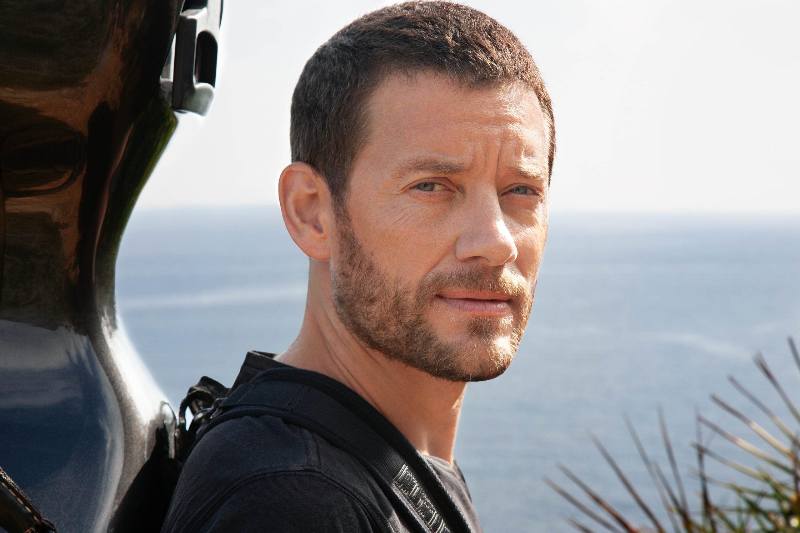 Músico Matthieu Saglio con el violonchelo en la espalda, con el cielo y el mar al fondo. Foto Ana Guimaras
